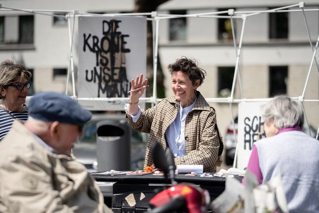 Die Künstlerin Tanja Krone sitzt umringt von Menschen auf einem öffentlichen Platz und winkt