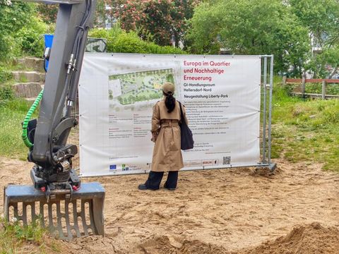Eine Baggerschaufel fängt an, in die Erde des Liberty Parks zu graben. Im Hintergrund ein Info-Plakat mit den Plänen des neuen Parks.