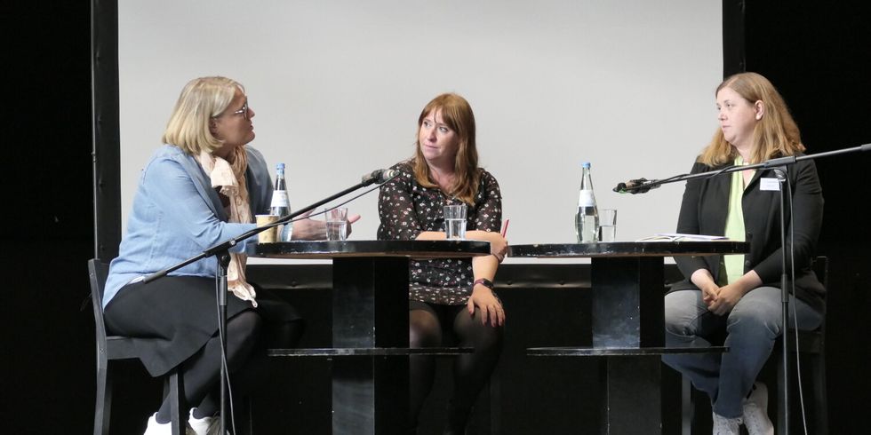 Drei Frauen sitzen auf einer Bühne zusammen an einem Tisch und reden miteinander.