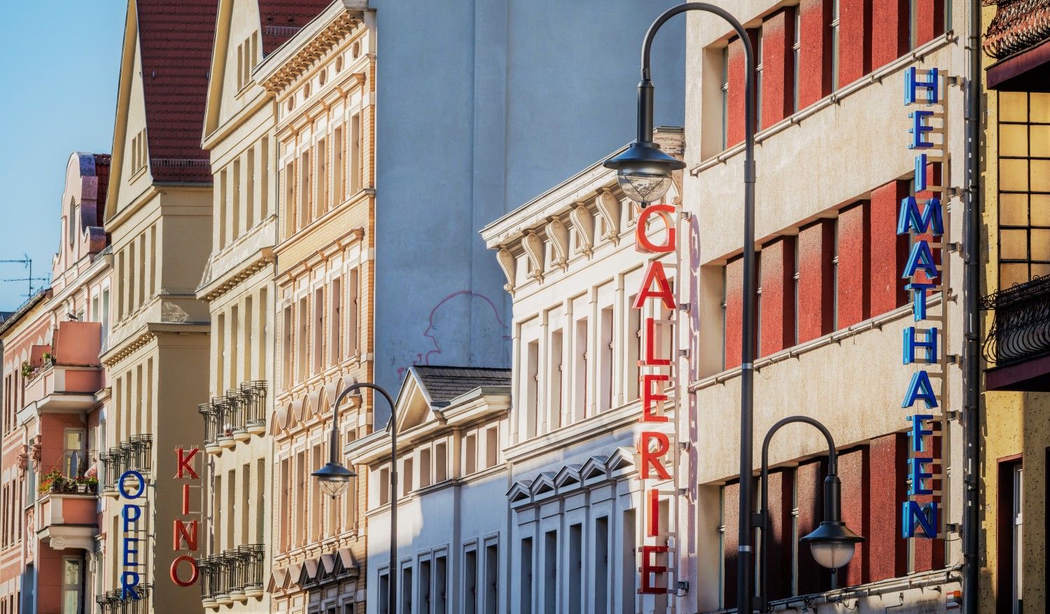 Häuserfassaden bei Tageslicht. Es sind die Schriften Heimthafen, Galerie, Oper und Kino zu sehen. Die Schriften sind senkrecht nach unten geschrieben und bestehen aus einzelnen Leuchtbuchstaben, die am Tag nicht an sind. Die Schriftzühe Heimathafen und Oper bestehen aus blauen Leuchtbuchstaben. Die Schriftzüge Galerie und Kino sind aus roten Leuchtbuchstaben zusammengesetzt