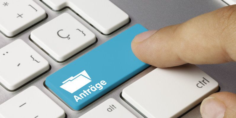 AUsschnitt einer Tastatur mit einer blau hervorgehobeben Taste auf der Anträge steht und von einem Finger berührt wird