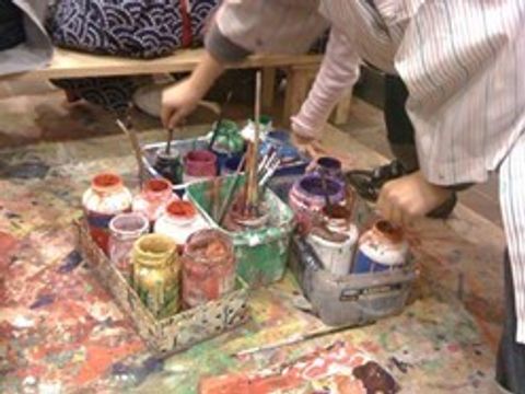 Viele offene Farbflaschen und Gläser stehen in Boxen auf einem bunt beklecksten Tisch. Es greifen Hände nach den vielzähligen Pinseln in den Behältnissen.