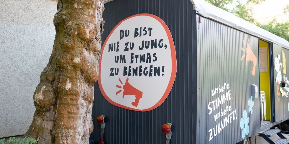Ein begehbarer Anhänger mit bunten Symbolen und dem Schriftzug "Du bist nie zu jung, um etwas zu bewegen!" steht mit offener Tür auf einem Platz.