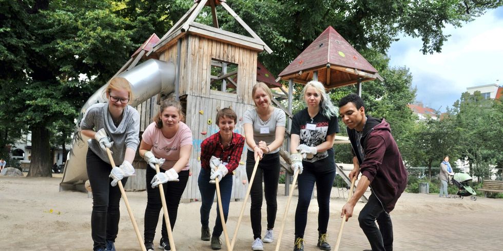 Pressefoto PULS-Camp 2023 - Sechs unterschiedliche Jugendliche kehren gemeisam Sand in die Fugen eines neu verlegten Pflasters auf einem Spielplatz