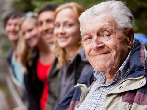 Ein älterer Mann sitzt zusammen mit einer Gruppe jüngerer Menschen und alle lachen Richtung Kamera