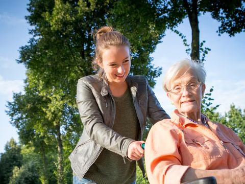 ein junge Frau schiebt eine ältere Dame im Rollstuhl durch einen Park