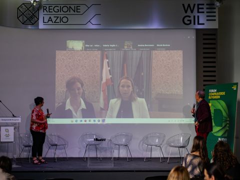 Auf einer Bühne stehen zwei Menschen, im Hintergrund ein Livestream mit Staatssekretärin Uleer und Referentin für Ernährung Weber