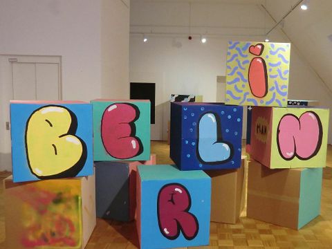 Große Würfel aus Karton mit bunten Grafiken bilden einen Stapel, unter anderem sind die Buchstaben "B, E, R L, I, N" zu sehen