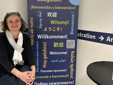 Vor einem Banner, auf dem "Willkommen!" in verschiedenen Sprachen steht, sitzt eine Frau.
