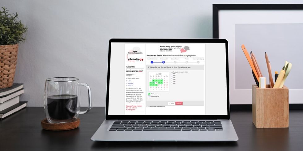 Laptop mit Online Terminierung vom Jobcenter Berlin Mitte, links daneben eine Tasse Kaffe und rechts ein Stiftehalter mit Stiften