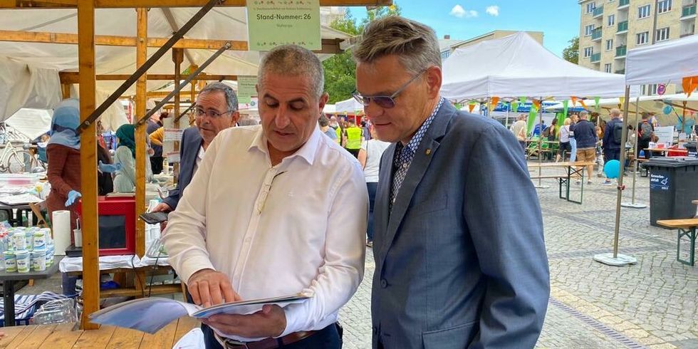 Zwei Männer stehen an einem Stand auf einem Festplatz und schauen zusammen in eine Broschüre.
