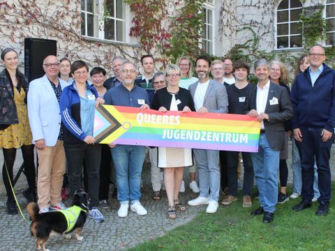 Mehrere Personen stehen zusammen vor einem Haus und schauen in die Kamera. Sie halten eine große LGBTIQ Flagge