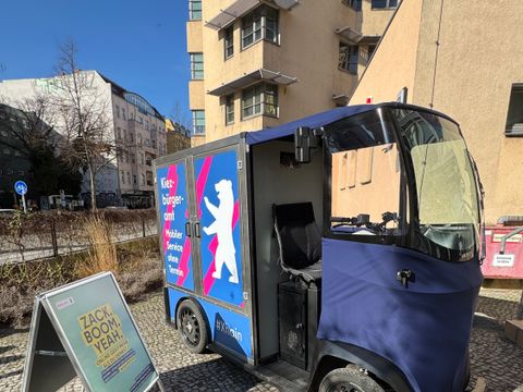 Lastenfahrrad mit Kofferaufbau mit blauem Werbeaufdruck und offenem Fahrersitz steht auf gepflastertem Gehweg neben einem aufgestellten Werbeschild