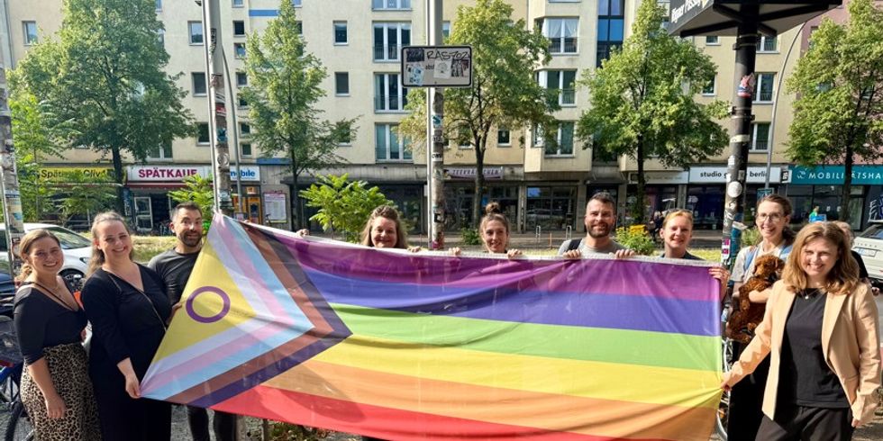 „Gemeinsam anders“ lautet das Motto des Bezirksamtes Friedrichshain-Kreuzberg und so darf auch die „Progress-Flag“ anders herum präsentiert werden: Bezirksbürgermeisterin Clara Hermann (re.) und Mitarbeiter*innen vor dem Rathaus Friedrichshain-Kreuzberg.