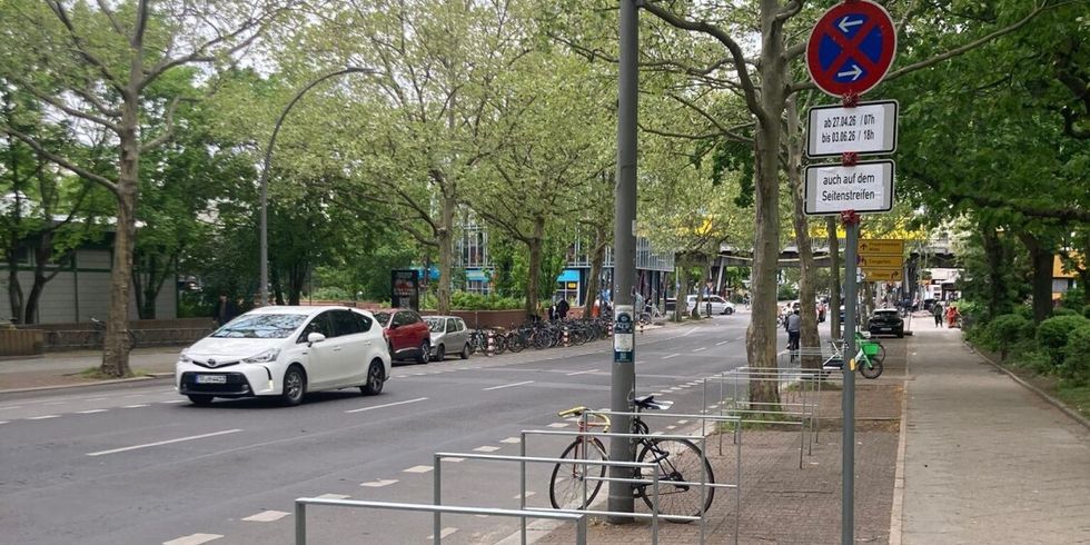 Mehrere silberne Radbügel auf einem gepflasterten Gehweg neben einer Straße mit parkenden Autos und Bäumen