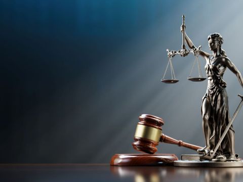 Eine weibliche Figur (genannt Justitia) in langem Gewand mit verbundenen Augen. In der einen Hand hält sie eine Waage, in der anderen ein gesenktes Schwert. Die Darstellung steht für Unparteilichkeit, Abwägung und Durchsetzung des Rechts. Daneben liegt ein brauner Richterhammer.