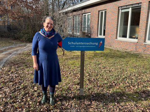 Eine Person mit blauem Kleid, gemustertem Schal und dunklen Stiefeln steht auf Laub und Gras neben einem Holzpfosten mit einem blauen Richtungsschild. Das Schild trägt den Schriftzug 'Schuluntersuchung Kinder- und Jugendgesundheitsdienst'. Im Hintergrund ist ein Backsteingebäude mit großen Fenstern und kahlen Bäumen zu sehen.