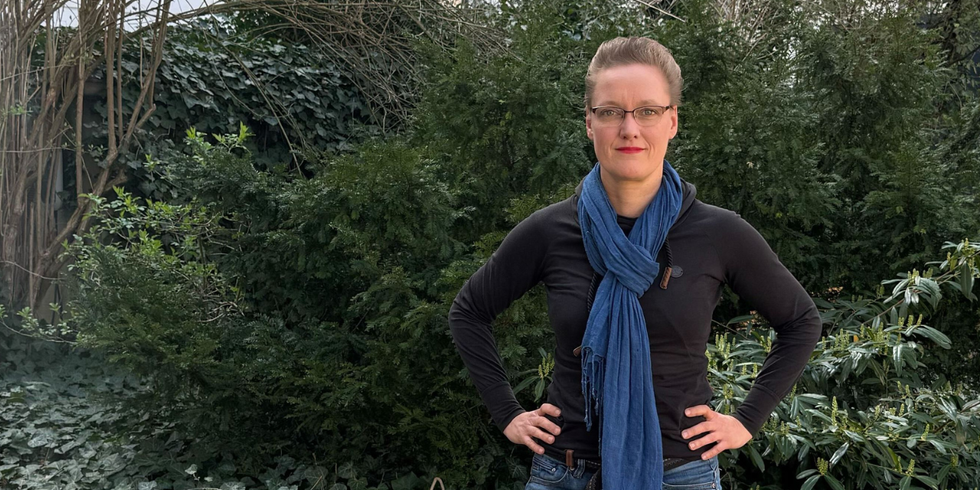 Romana Wittmer, Beauftragte für Gute Arbeit im Bezirksamt Friedrichshain-Kreuzberg, steht vor einer begrünten Fläche.