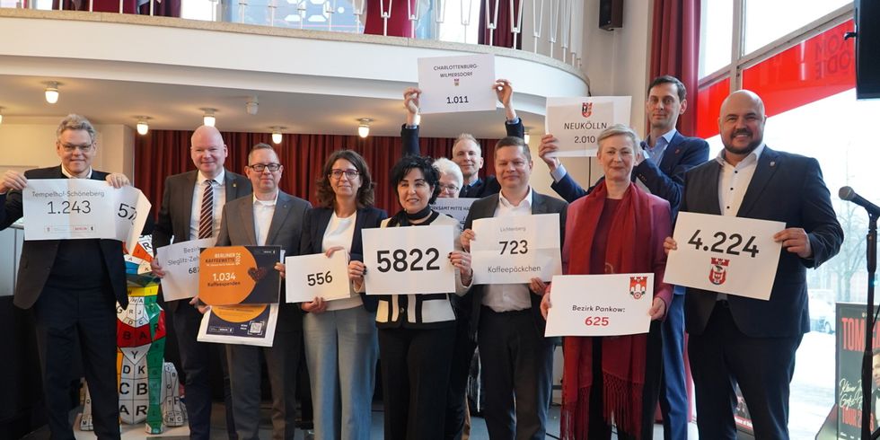 Bezirksbürgermeisterinnen und Bezirksbürgermeister präsentieren die Ergebnisse der Kaffeewette 2026 und halten Schilder hoch mit der Anzahl der gesammelten Kaffeepäckchen.