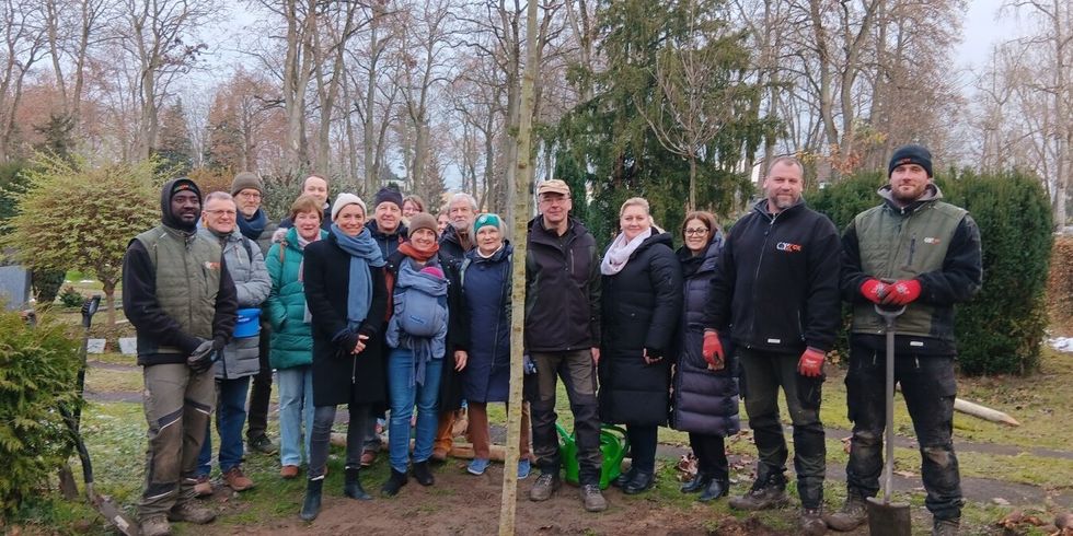 Mehrere Personen stehen zusammen auf einem Friedhof. In der Mitte steht ein neu gepflanzter Baum.