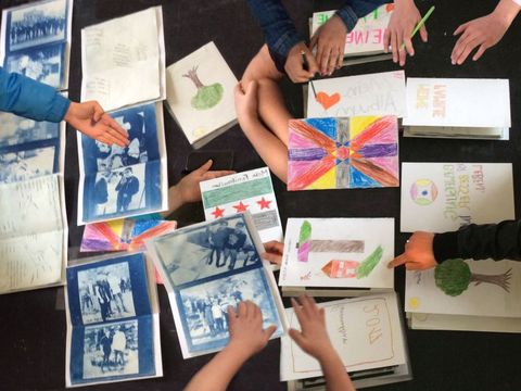 Auf einem Tisch liegen einige aufgeschlagene, selbst hergestellte Leporello-Fotoalben mit bläulichen Fotos, genannt Cyanotypien. Daneben befinden sich einige geschlossene Alben mit Umschlägen, die mit farbigen Motiven - wie Bäumen oder Häusern - in Buntstift gestaltet wurden.