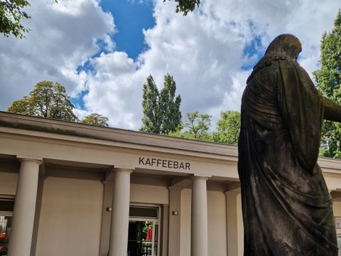 Eingang zur Kaffeebar mit Säulengang, im Vordergrund Anschnitt einer Statue