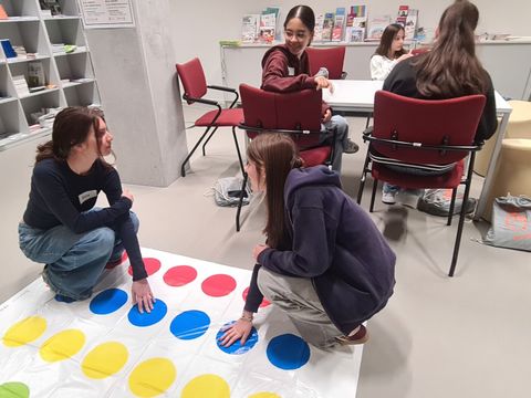 Im Vordergrund spielen zwei Mädchen auf einer Twister-Matte, die mit großen, farbigen Kreisen in Rot, Gelb, Grün und Blau bedruckt ist. Beide knien auf dem Boden und berühren mit Händen und Füßen verschiedene Farbfelder. Im Hintergrund sitzen weitere Jugendliche an einem Tisch, unterhalten sich und beschäftigen sich mit Materialien. Die Umgebung wirkt wie ein Aufenthaltsbereich mit Regalen voller Broschüren und Informationsmaterialien. Die Szene findet im Rahmen des Girls' Day im Jobcenter Friedrichshain-Kreuzberg statt.