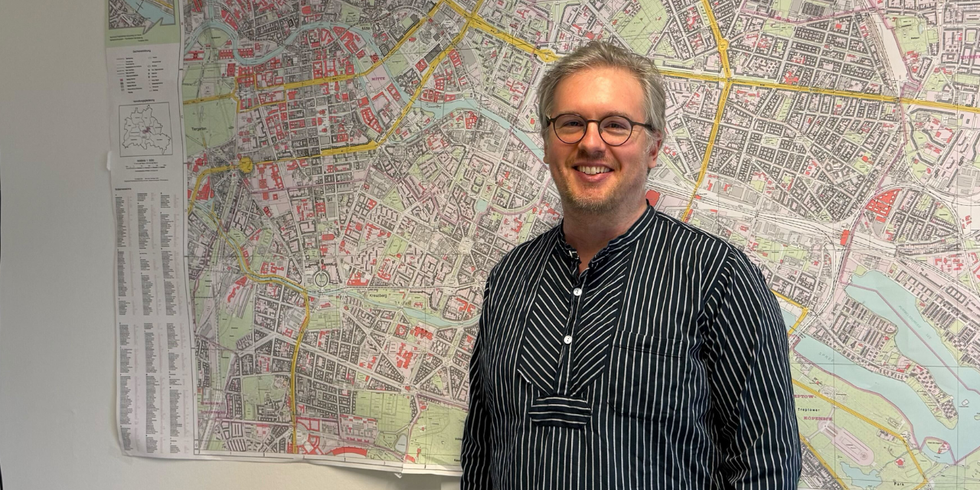 Aaron Schaar steht mit neutralem Gesichtsausdruck und gestreiftem Hemd vor einer großformatigen Stadtkarte an der Wand.