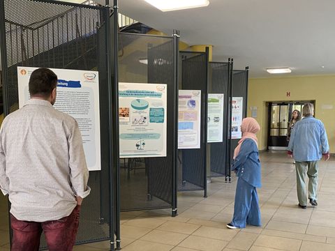 Vor den Ausstellungswänden der Ausstellung über gendersensible Medizin der VHS Neukölln stehen drei Personen an verschiedenen Stellen und schauen sich die Poster an den Stellwänden an.