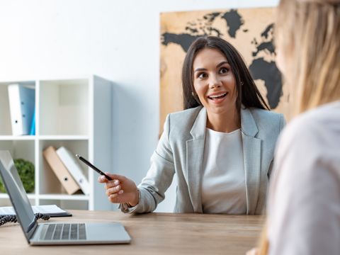 Lächelnde Frau am Tisch im Gespräch mit anderer Frau zeigt auf auf einen Laptop