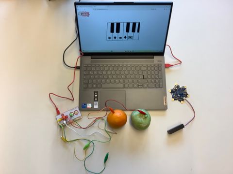 Laptop mit Obst davor - alles ist über Kabel miteinander verbunden. Auf Bildschirm ist Klaviatur zu sehen