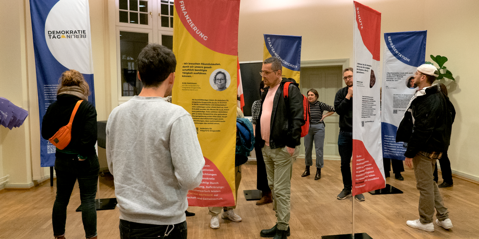 Menschen stehen in einem Raum und schauen auf Fahnen mit Informationen.