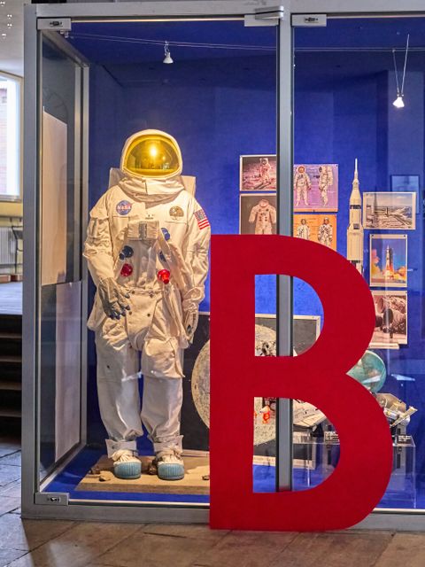Ein Raumfahranzug steht in einem Schaufenster mit Bildern und Modellen aus der Raumfahrt. Vor dem Schaufenster ist ein großes rotes B.