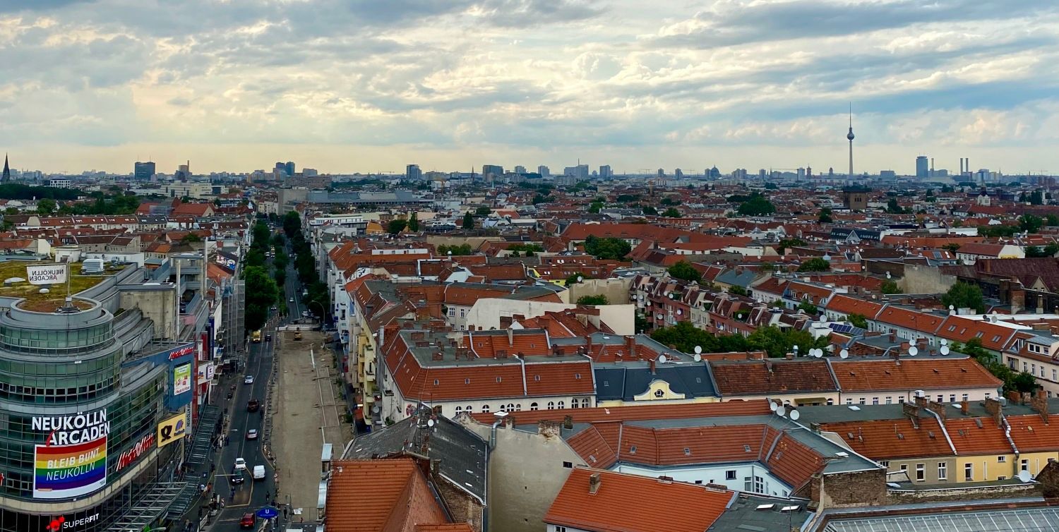 Bild von oben. Blick auf die Karl-Marx-Straße in Richtung Fernsehturm. Man sieht viele Häuser von oben. Man sieht die Dächer. Die meisten haben Dächer mit roten Dachschindeln. Der Himmel ist wolkenverhangen aber durch die Wolken kann man die Sonne erahnen