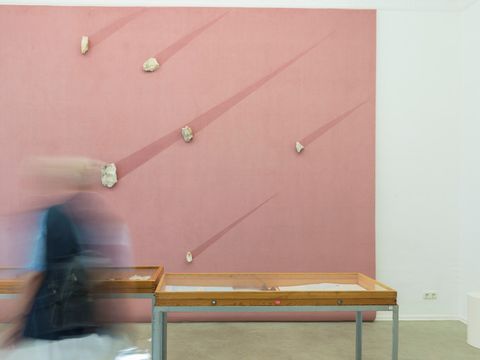 Im Hintergrund ein rosanes Bild mit hellen aufgeklebten mehreren größeren Steinen. Sie ziehen einen dunklen rosa Schweif hinter sich, wie eine Sternschnuppe. Davor zwei Tische. Es ist eine Kunstinstallation. Auf der linken Bildseite ist eine Frau mit einem dunklen Oberteil zu sehen. Sie ist gerade in Bewegung und sehr verschwommen aufgenommen.