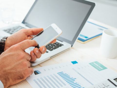 Ein Mann sitzt vor seinem Laptop und stellt Berechnungen an. Man sieht lediglich seine Hände, die ein Smartphone bedienen.
