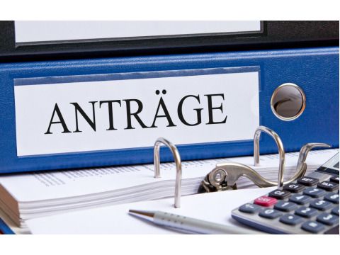 Auf einem geöffneten Aktenordner liegen ein Taschenrechner, ein Stift, mehrere Geldscheine und weitere Ordner mit der Aufschrift 'Anträge'