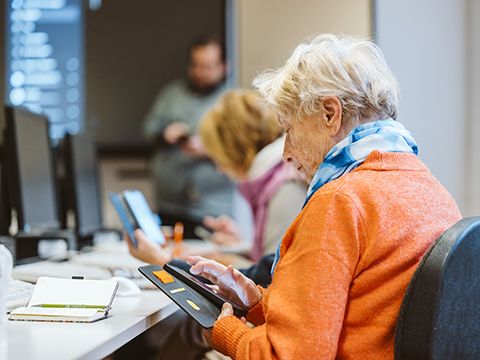 eine ältere Frau mit dem Smartphone in der Hand; im Hintergrund der Kursleiter