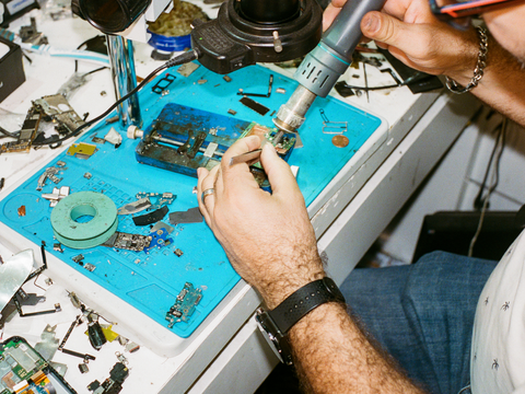 Eine Person repariert elektronische Geräte auf einer Werkbank, umgeben von Werkzeugen und Ersatzteilen. Sie nutzt einen Lötkolben, um kleine Bauteile zu bearbeiten. Das Bild zeigt die Bedeutung von Reparatur und Wiederverwendung im Sinne der Zero-Waste-Bewegung und fördert eine nachhaltige Nutzung von Elektrogeräten.