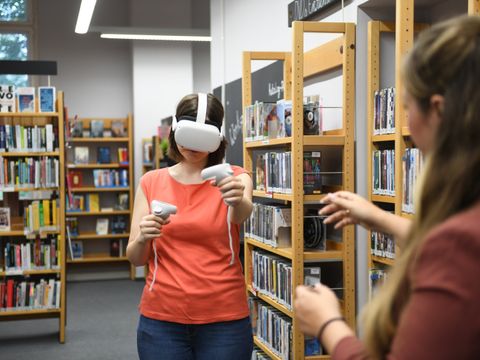 Person mit VR-Brille und Controllern in der Kurt-Tucholsky-Bibliothek, begleitet von einer weiteren Person