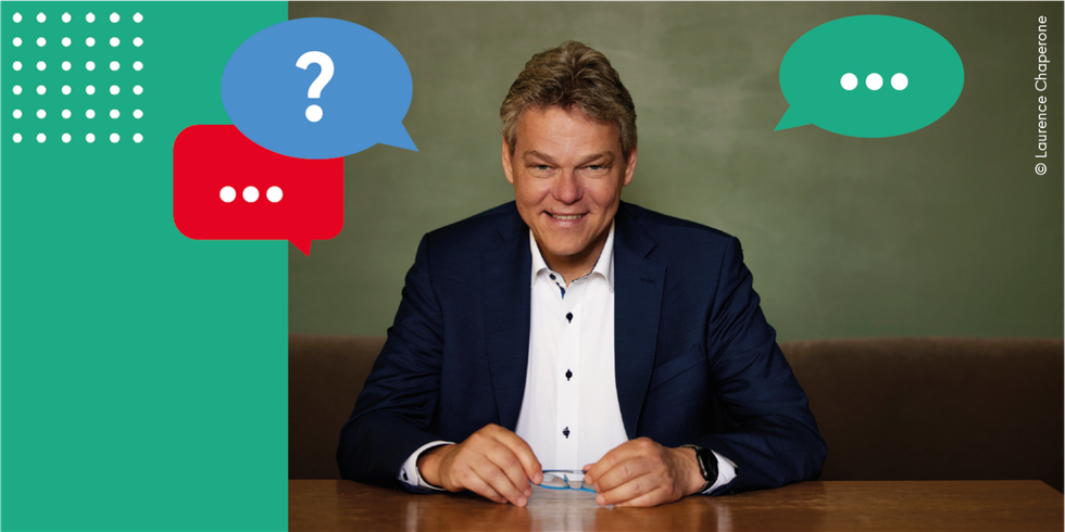 Ein Mann sitzt an einem Tisch und lächelt in die Kamera. Von ihm ausgehend sind bunte Sprechblasen eingefügt.