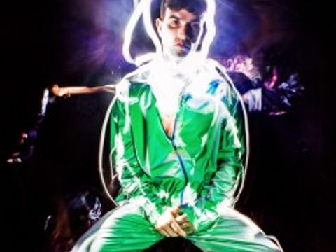 Eine sitzende Person in einem glänzenden grünen Trainingsanzug und weißen Sneakers vor dunklem Hintergrund. Um ihren Kopf und Oberkörper zeichnen sich helle Lichtspuren, die wie leuchtende Linien wirken und einen starken Kontrast zur Umgebung bilden. Die Szene hat eine künstlerische, experimentelle Lichtinszenierung.