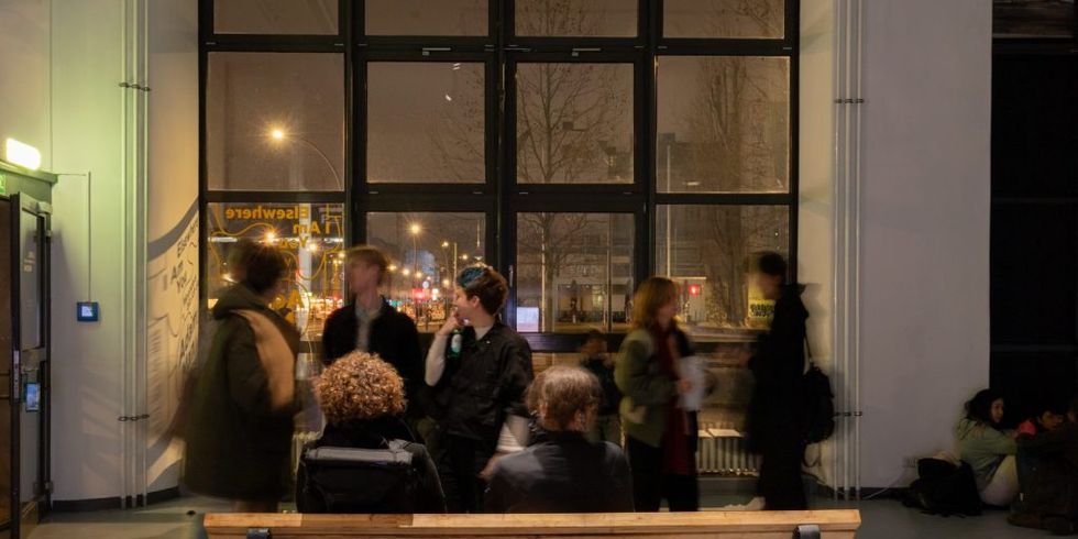 Menschen auf einer Parkbank, dahinter Menschen im Gespräch vor einem großen Fenster