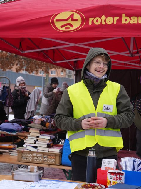 Jonas vom Raum für Beteiligung steht lächelnd vor dem Stand am Kieztag - vor ihm liegen Broschüren, Notizbücher und Süßigkeiten zum Verteilen.