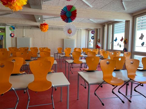 Mehrere orangefarbene Stühle stehen in gleichmäßigen Reihen an weißen Tischen in einem Klassenraum. Die Stühle sind aus Kunststoff und haben eine charakteristische Öffnung in der Rückenlehne. An den Fenstern und Wänden hängen bunte Papierrosetten und Dekorationen. Der Boden ist rot, die Wände und Decke bestehen aus hellem Holz. Große Fenster lassen viel Tageslicht herein.