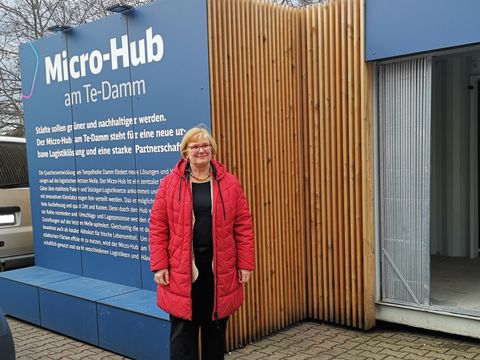 Frau steht vor einem eckigen Bauwerk mit Eingang mit der Aufschrift Micro-Hub am Te-Damm