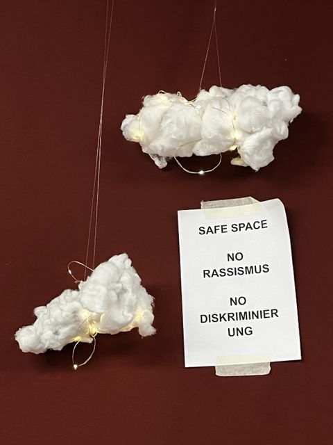 Wolkenlampen hängen vor braunem Hintergund. Daneben ein Zettel mit der Aufschrift "Safe Space. No Rassismus. No Diskriminierung."