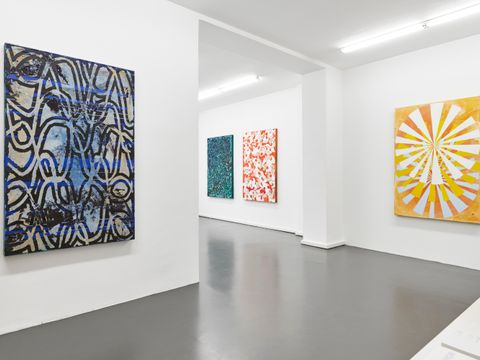 Das Bild zeigt eine Galerie. Auf der linken Seite ist eine weiße Wand. Darauf hängt ein quadratisches Bild. Es ist in blau und weiß gehalten. Es sind mehrere verschlungene helle Linien zu sehen. Auf der rechten Seite ein weiteres quadratisches Bild. Auf einem gelben HIntergrund sind strahlenförmig weiße Streifen zu sehen. Links neben dem Bild ist ein Vorsprung. Dahinter eine weitere weiße Wand, auf der zwei weitere quadratische Bilder angebracht sind.