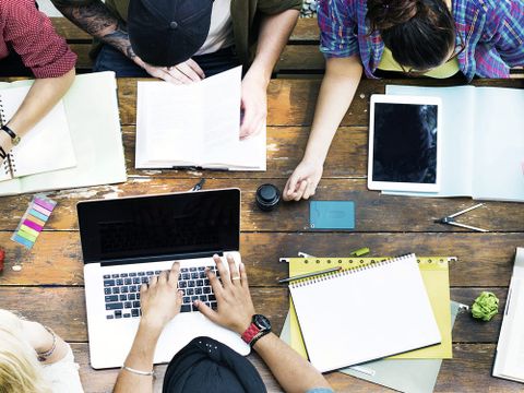 Junge Menschen an einem Holztisch mit Papieren, Stiften, Lineal, Zirkel und Laptops