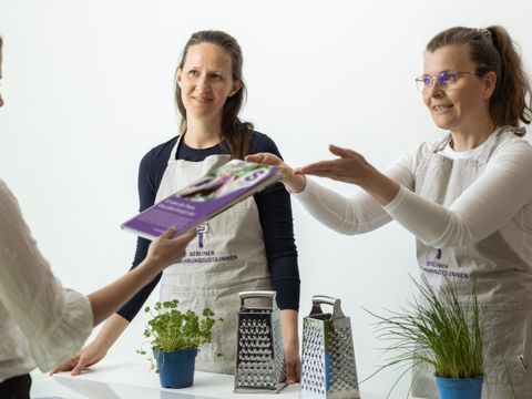 Zwei Frauen mit Kochschürzen stehen hinter einem Tisch, eine der Frauen reicht einer dritten Frau vor dem Tisch eine Zeitschrift